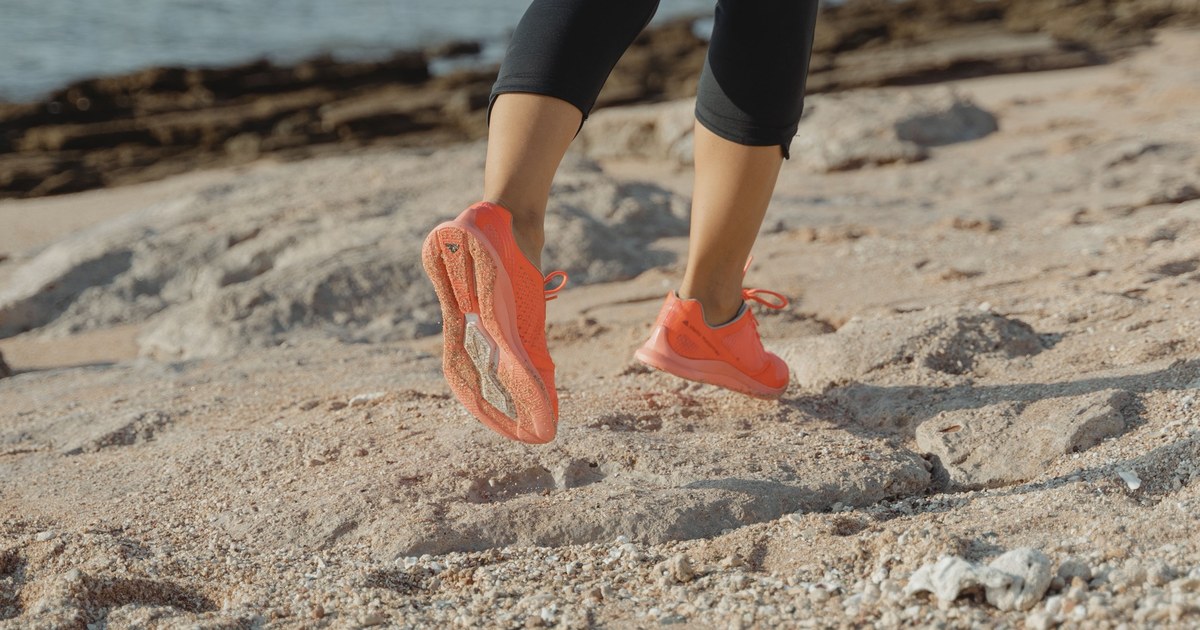 Pies de una corredora con zapatillas naranjas sobre rocas y arena junto al mar